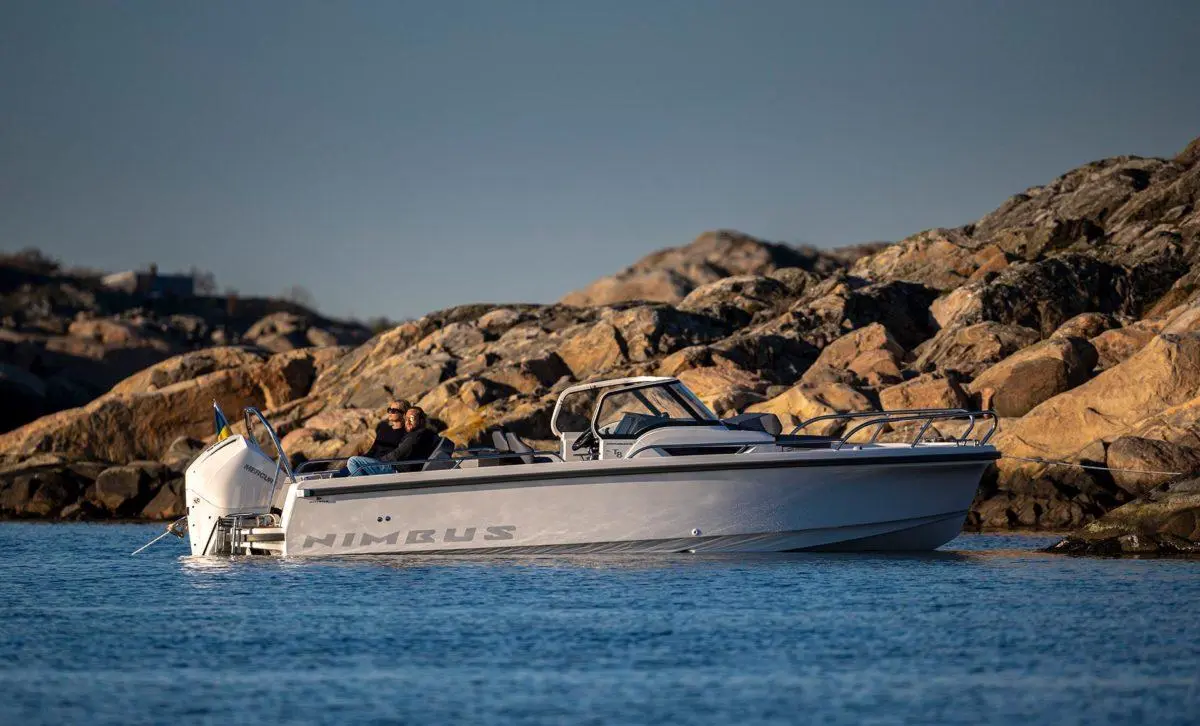 Two people relax on a Nimbus boat near rocky coastline under clear skies, showcasing marine leisure and exploration.